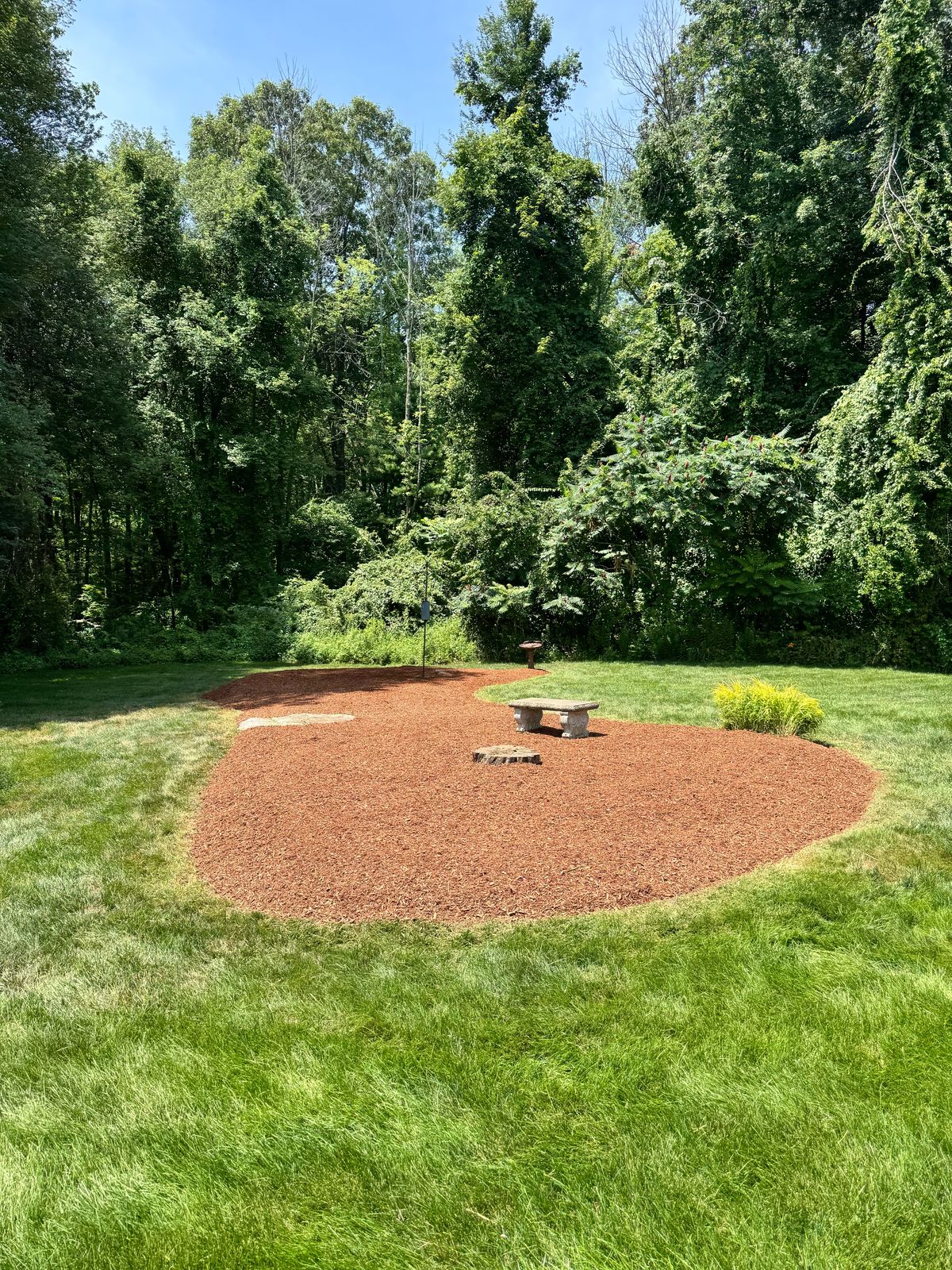 Large red mulch bed with a stone bench at the edge of a lawn